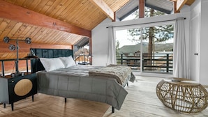 Un dormitorio con una cama grande, cabecera de madera, mesita de noche con lámpara y vistas al exterior a través de un ventanal corredero.