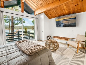 A bedroom with a large bed, a wooden bench, a wicker stool, and a view of a lake and mountains through a glass door. - Big Bear Lake View (Big Bear Lake)
