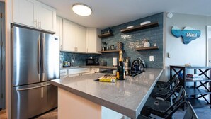A modern kitchen with a stainless steel refrigerator, a built-in oven, and a granite countertop. There are wooden shelves above the counter and a decorative wall piece.