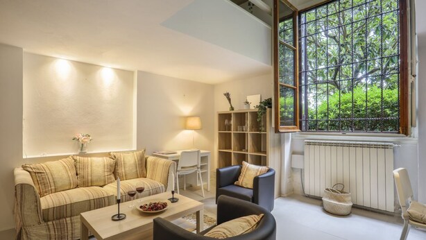 A living room with a sofa, a coffee table, and a chair. There is a window with a view of greenery outside. - Gelsomino - Sweet Dreams in Florence (Firenze)
