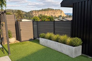 A fenced backyard with artificial grass, a concrete planter with plants, and a view of a mountain. - Grand Oasis Kelowna, Resort-Style Pool, Hot Tub, Sauna 5 Min to Downtown (Kelowna)