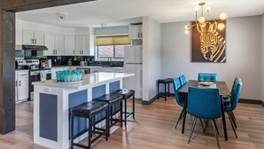 Een moderne keuken met witte kasten, een centraal eiland met krukken, en een eetgedeelte met blauwe stoelen.