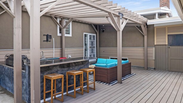 Een terras buiten met een houten dek, een bar met krukken, en een zithoek met kussens.