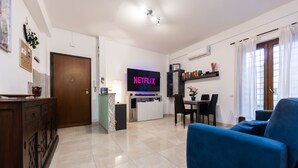 Een moderne woonkamer met een blauwe bank, een televisie waarop 'NETFLIX' staat, een eethoek en een keuken.