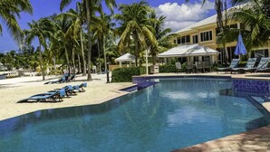 A pool area with lounge chairs and palm trees, surrounded by a building with a tiled pool edge. - Stunning beachfront condo. Relax, Enjoy, Sunset, RumPoint, Pool & Heated Jacuzzi (Grand Cayman)