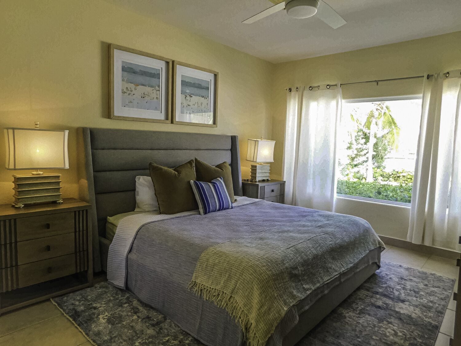 A bedroom with a large bed, two bedside tables with lamps, a ceiling fan, and a window with curtains.