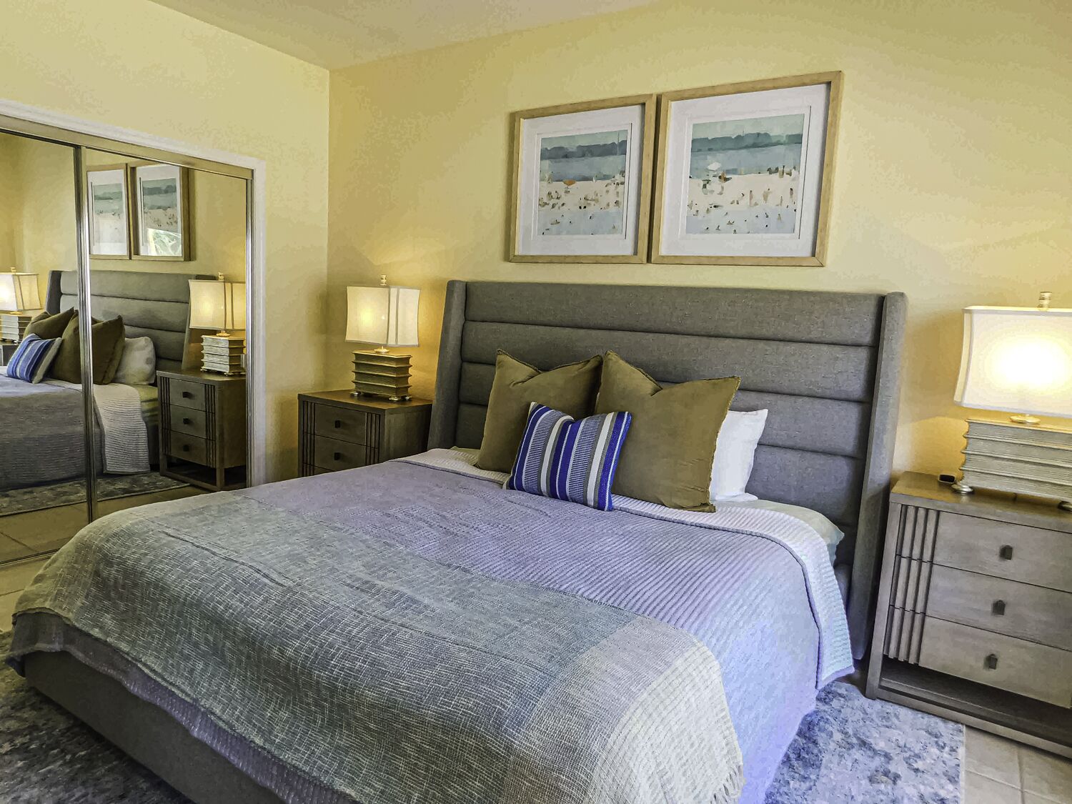 A neatly arranged bedroom with a large bed, a bedside table with a lamp, a nightstand, a mirror, and two framed pictures on the wall.