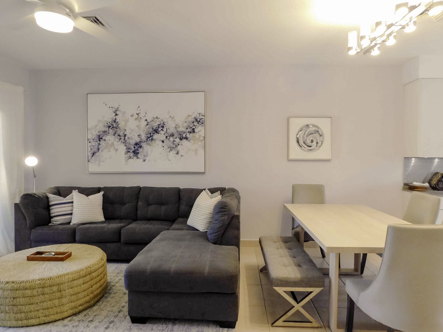 Un salon moderne avec un canapé d’angle gris, un pouf rond et une table à manger avec des chaises. Les murs sont ornés de tableaux abstraits.