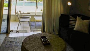A room with a view of a pool and palm trees, a round ottoman with a phone, a sofa, and a lamp. - Stunning beachfront condo. Relax, Enjoy, Sunset, RumPoint, Pool & Heated Jacuzzi (Grand Cayman)