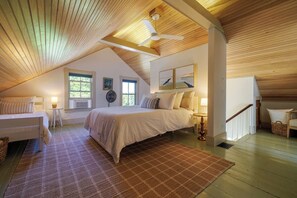 A bedroom with a sloped ceiling, a bed, a standing fan, and a window.