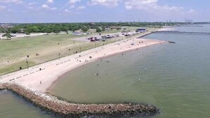 An aerial view of a coastal area with a sandy beach, a rocky breakwater, and a distant pier. - Cozy Duplex Close to Hobby, Lagoon, NASA, Beaches! (Pasadena)