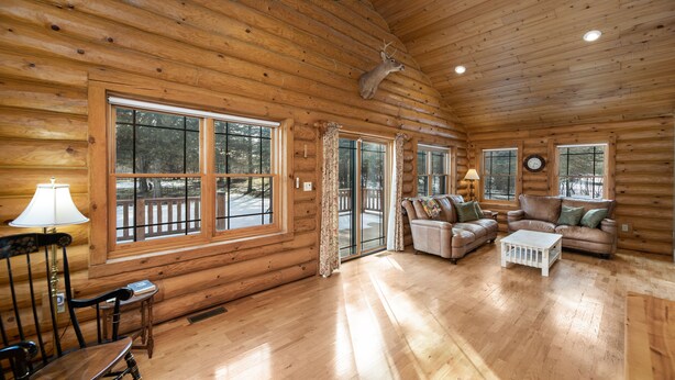 A log cabin interior with wooden walls, large windows, a fireplace, and a ceiling-mounted deer head.
