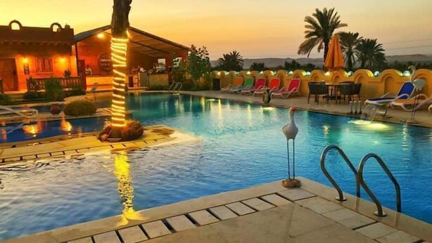 A swimming pool with lounge chairs and a palm tree, surrounded by a building with a warm glow. - Tunis Pyramids Hotel (Yusuf as-Sidiq)