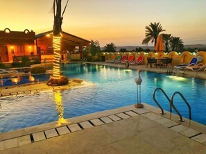 A swimming pool with lounge chairs and a palm tree, surrounded by a building with a warm glow. - Tunis Pyramids Hotel (Yusuf as-Sidiq)