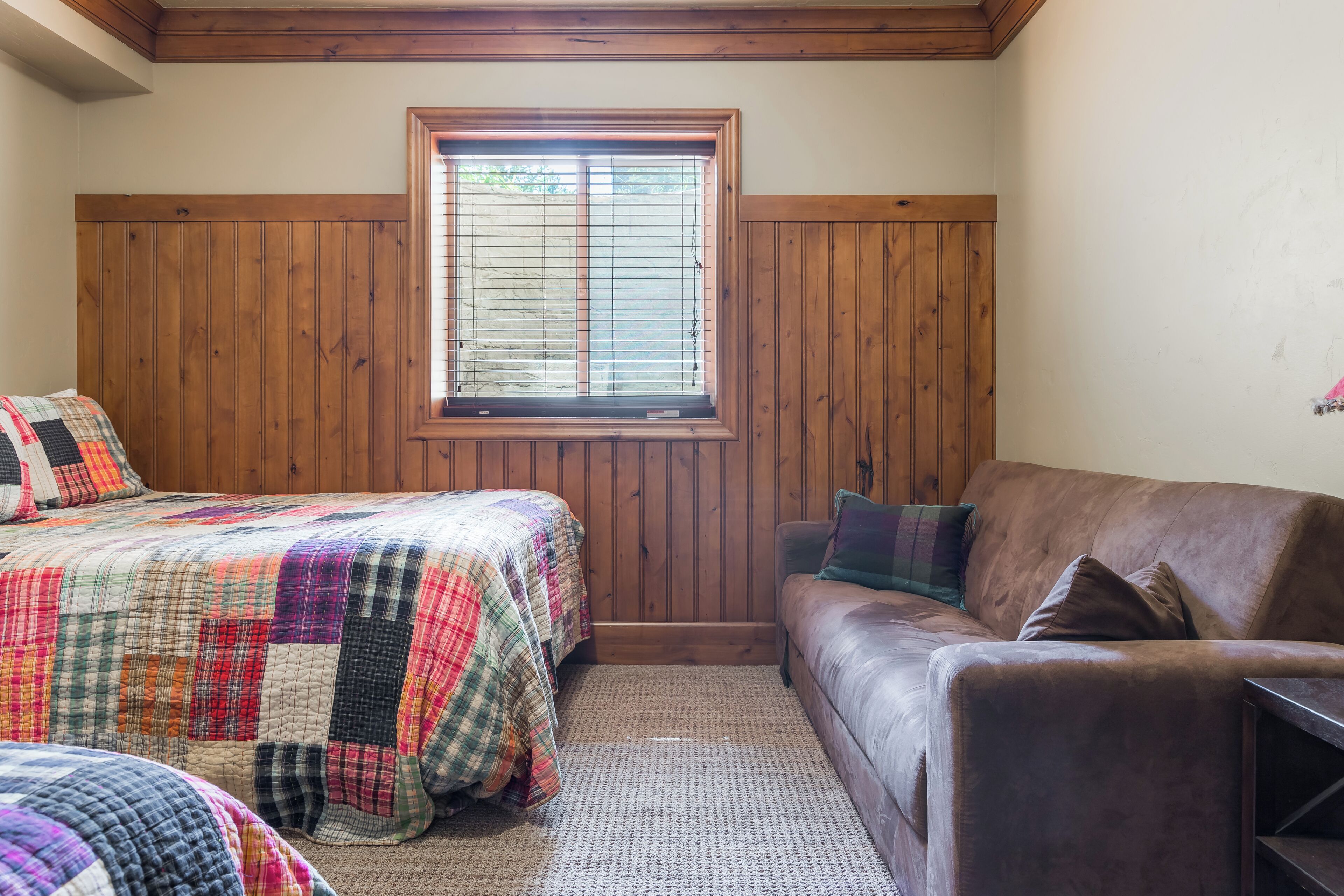 Un dormitorio con pared de paneles de madera, una ventana con persianas, una cama con colcha colorida, un sofá de cuero marrón y una mesita.