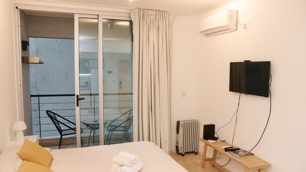 A bedroom with a bed, a TV, a small table, a radiator, and a balcony with a chair. - Silencioso Studio en Palermo Hollywood (Buenos Aires)