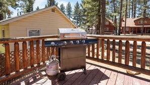Een barbecue op een terras met houten reling, een propaanfles en een gebouw op de achtergrond.
