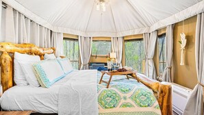 A cozy, tent-style bedroom with a large bed, wooden headboard, and a view of the forest through the windows.
