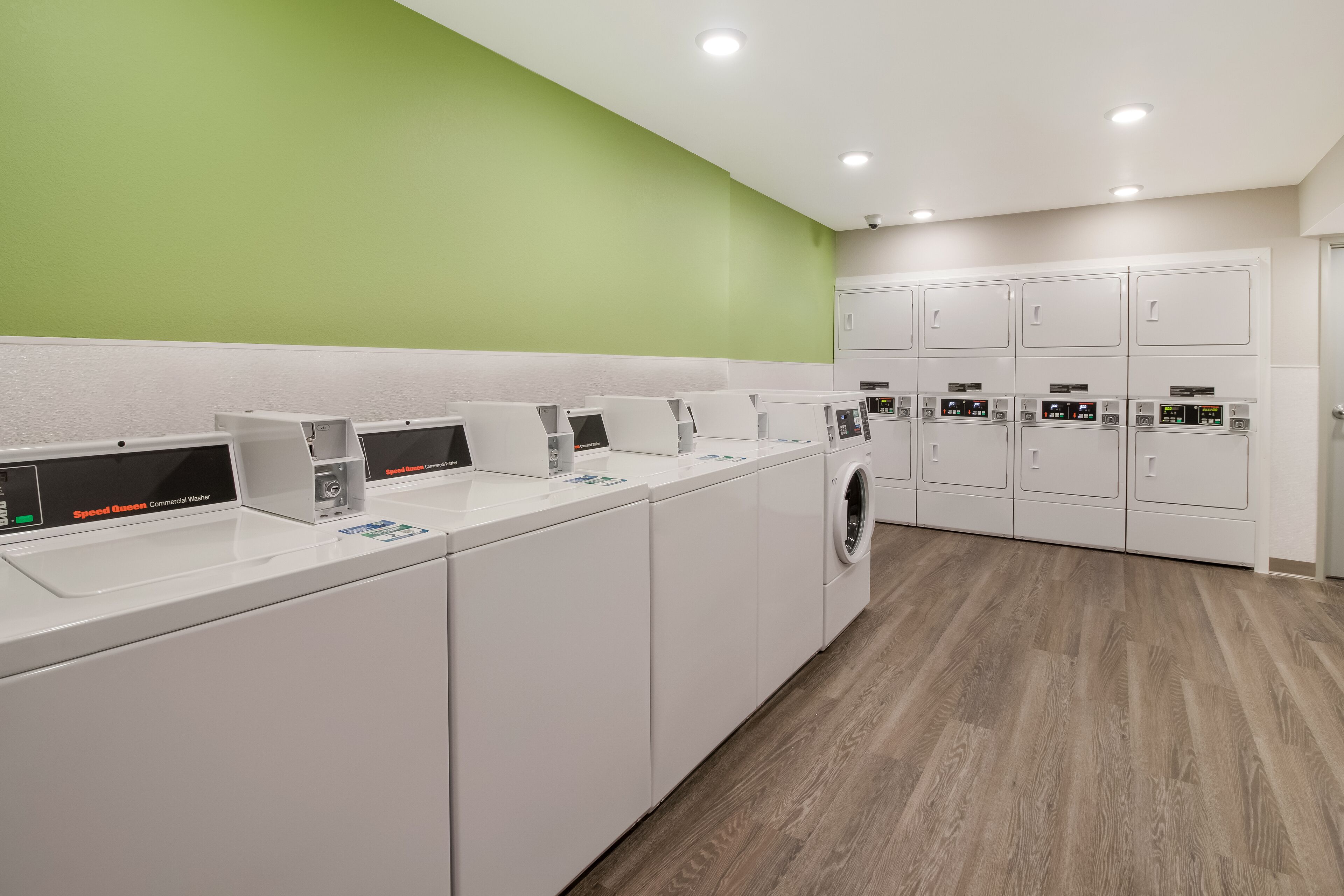 A modern laundry facility with multiple washing machines and dryers, a green wall, and a wooden floor.