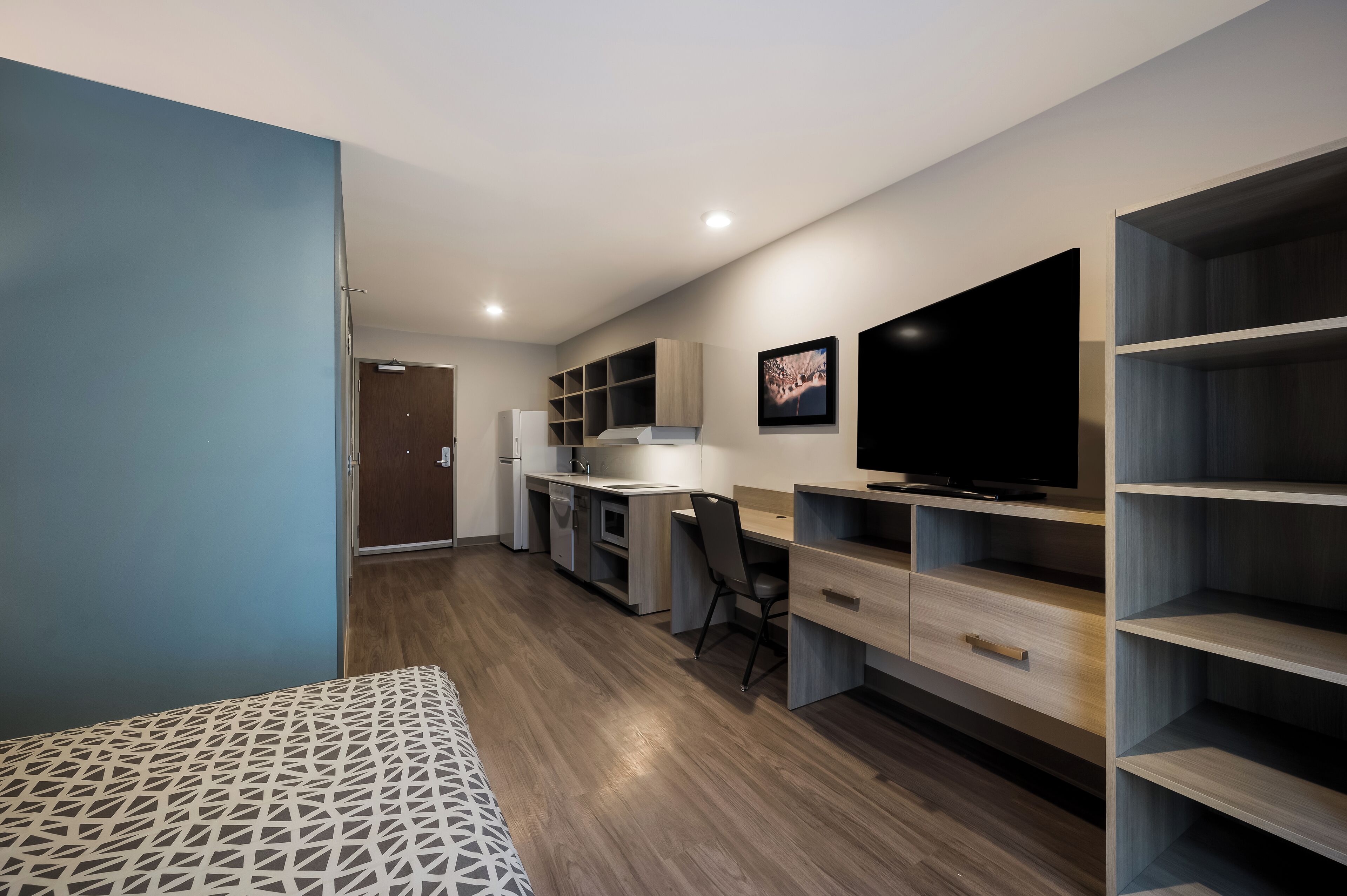 A modern, compact room with a kitchenette, a bed, a desk, and a TV.