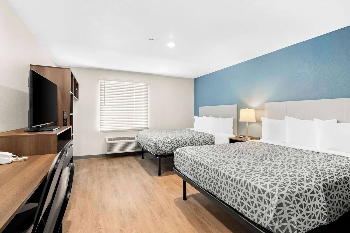 A hotel room with a large bed, a smaller bed, a desk with a TV, and a window with blinds.