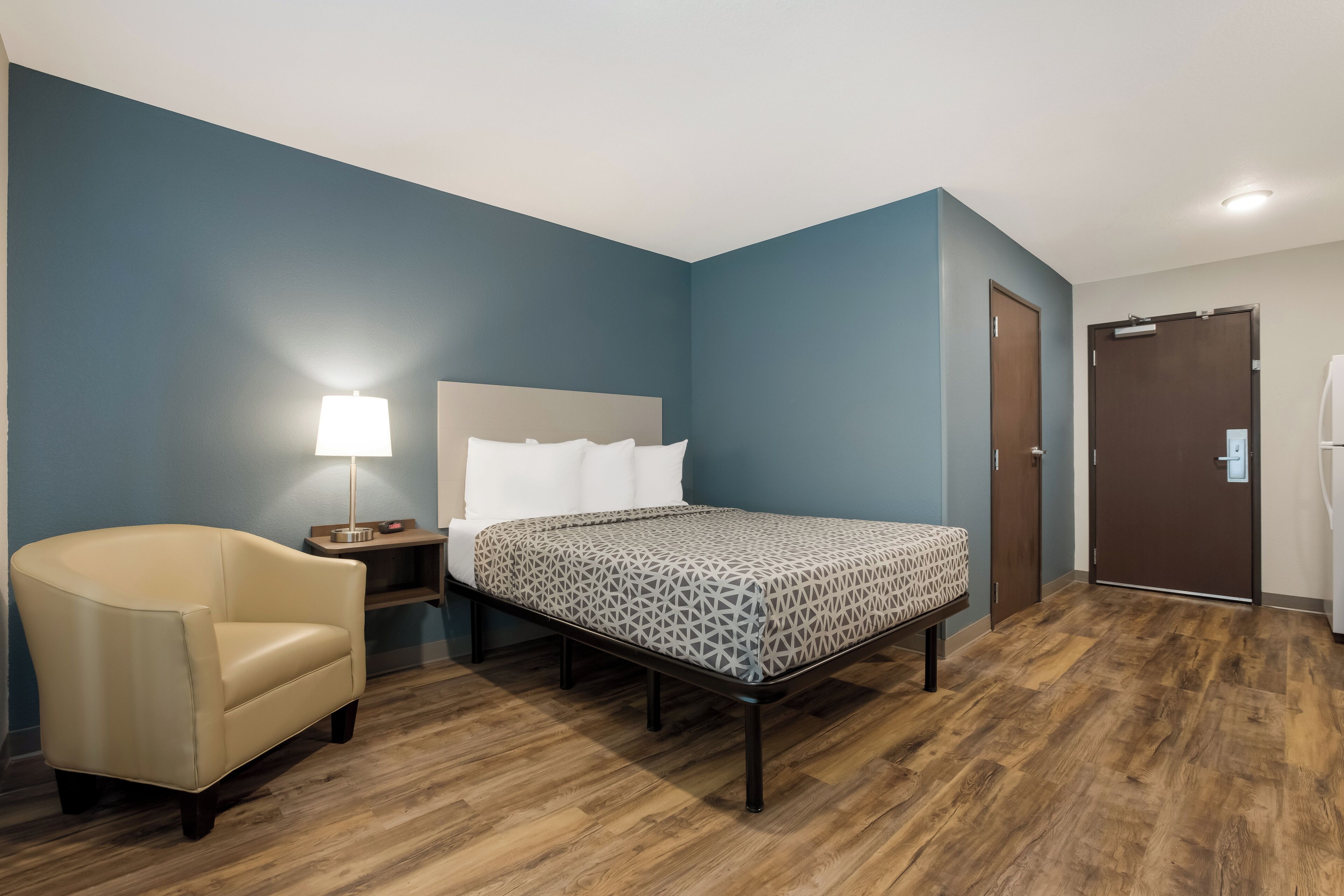 Una habitación de hotel con cama, sillón, mesita de noche, lámpara y puerta.