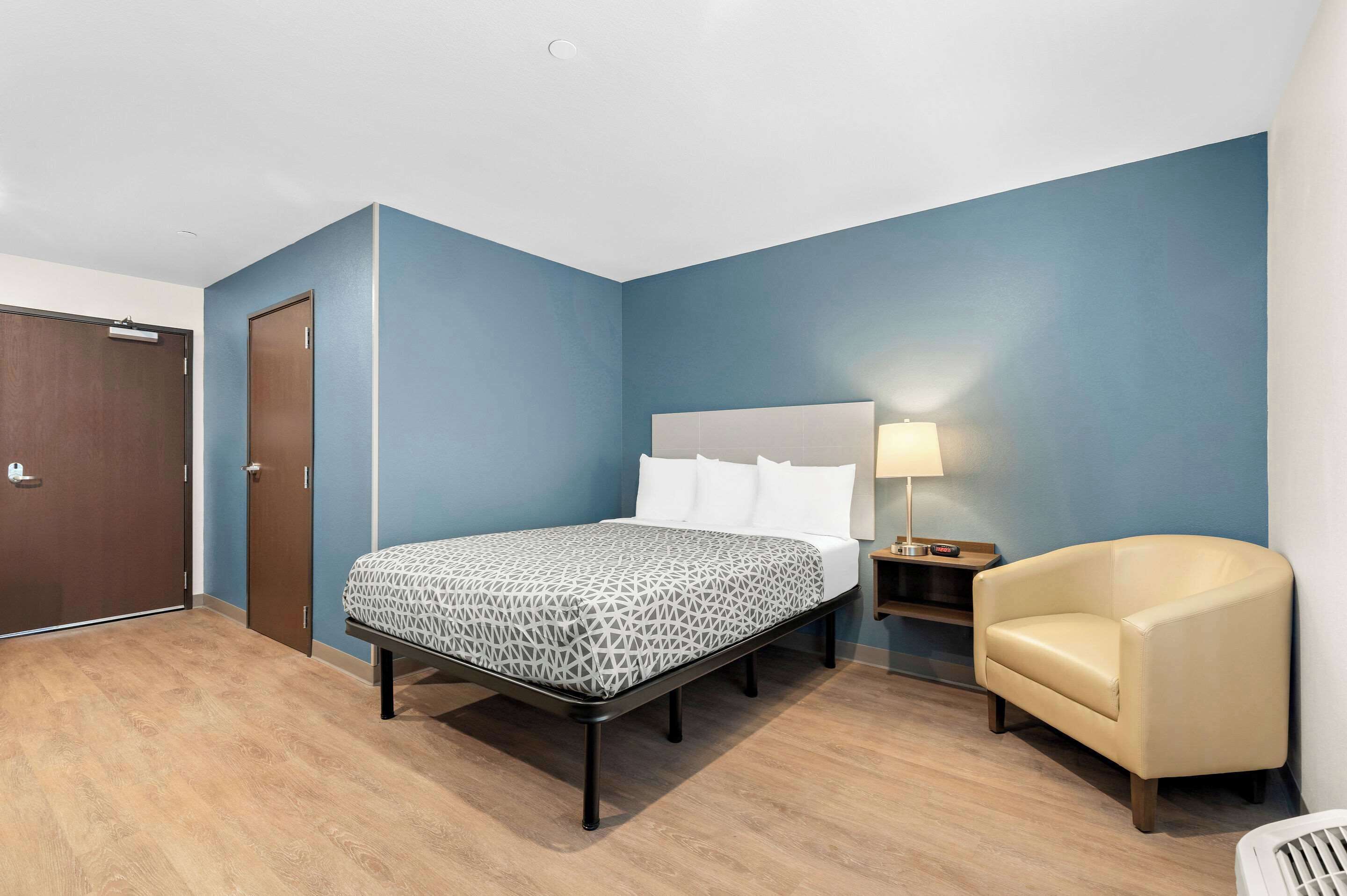 A hotel room with a bed, a chair, a nightstand with a lamp, and a door.