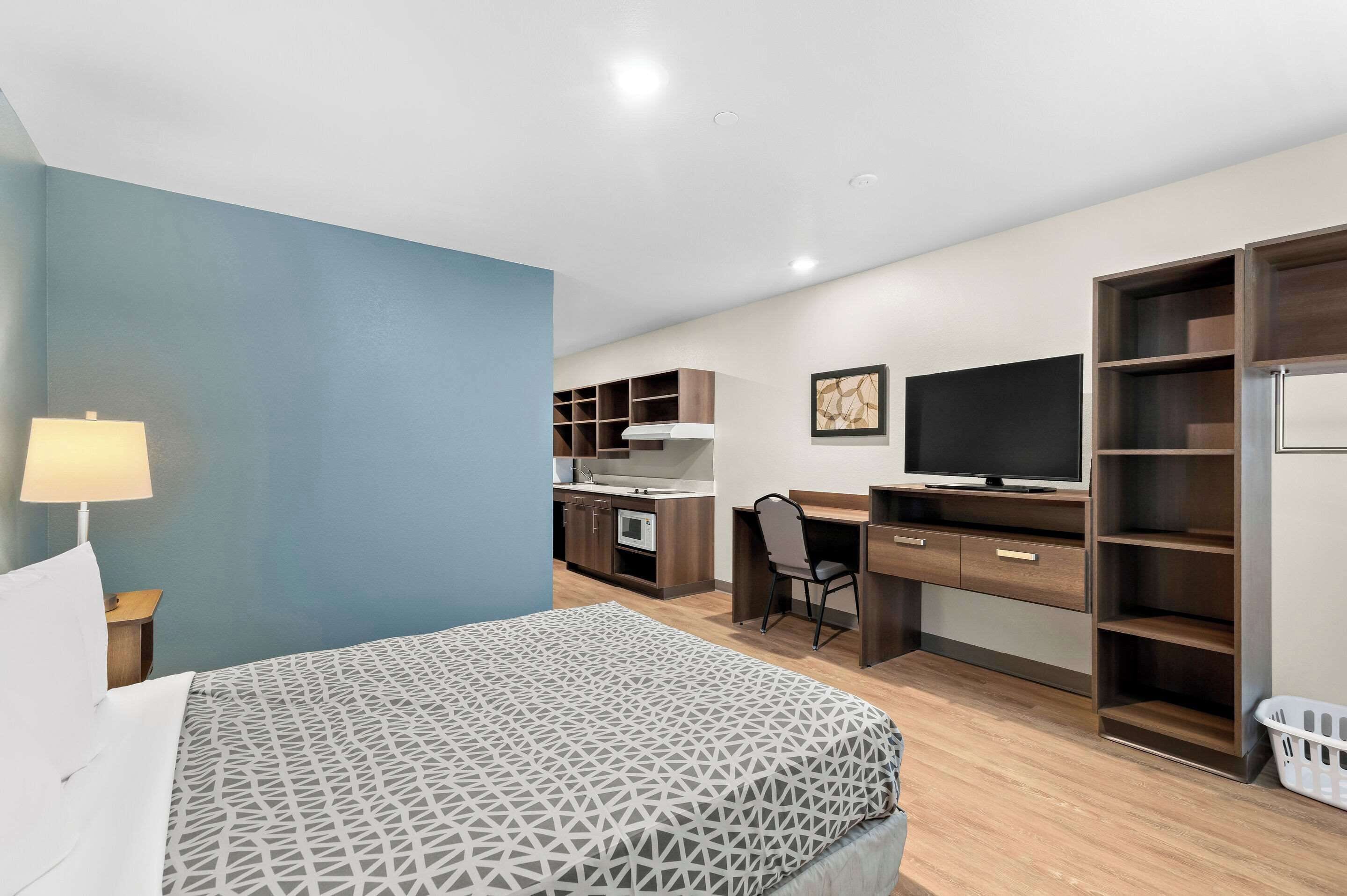 A modern hotel room with a bed, desk, kitchenette, and a TV.