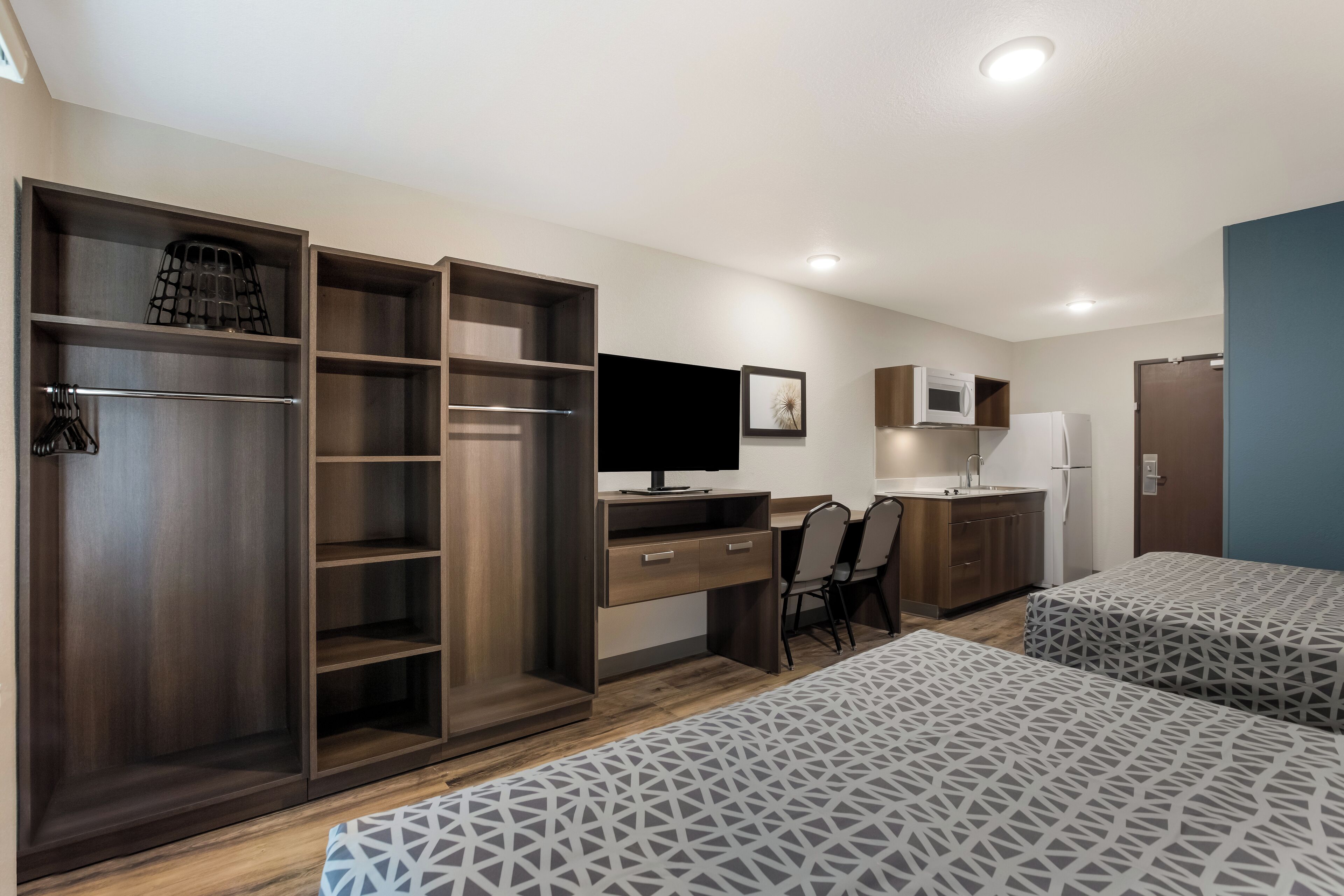 A compact room with a bed, a desk, a chair, a wardrobe, a TV, and a kitchenette.