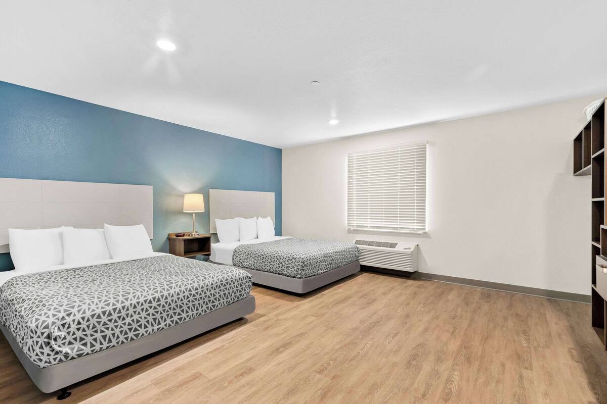 A hotel room with two beds, a nightstand, a lamp, a window with blinds, and a built-in wardrobe.