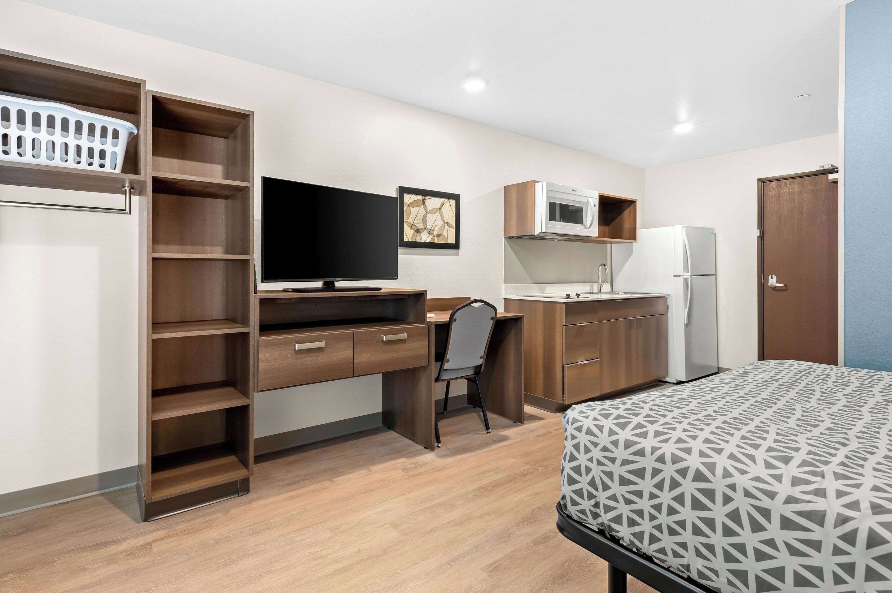 Habitación pequeña con cama, escritorio y silla, televisor, microondas, refrigerador y zona de cocina.