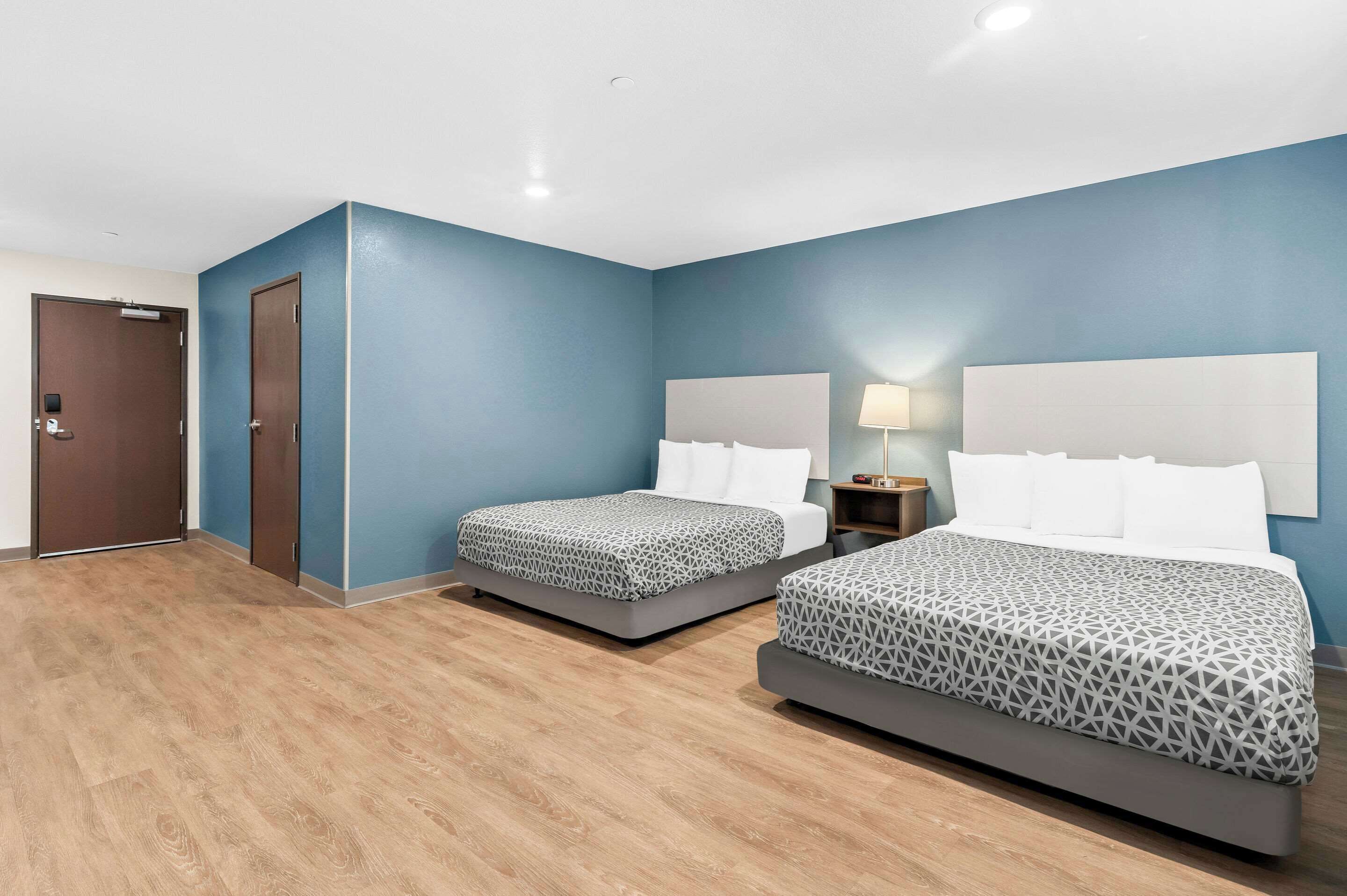 A hotel room with two beds, a wooden floor, a door, and a nightstand with a lamp.
