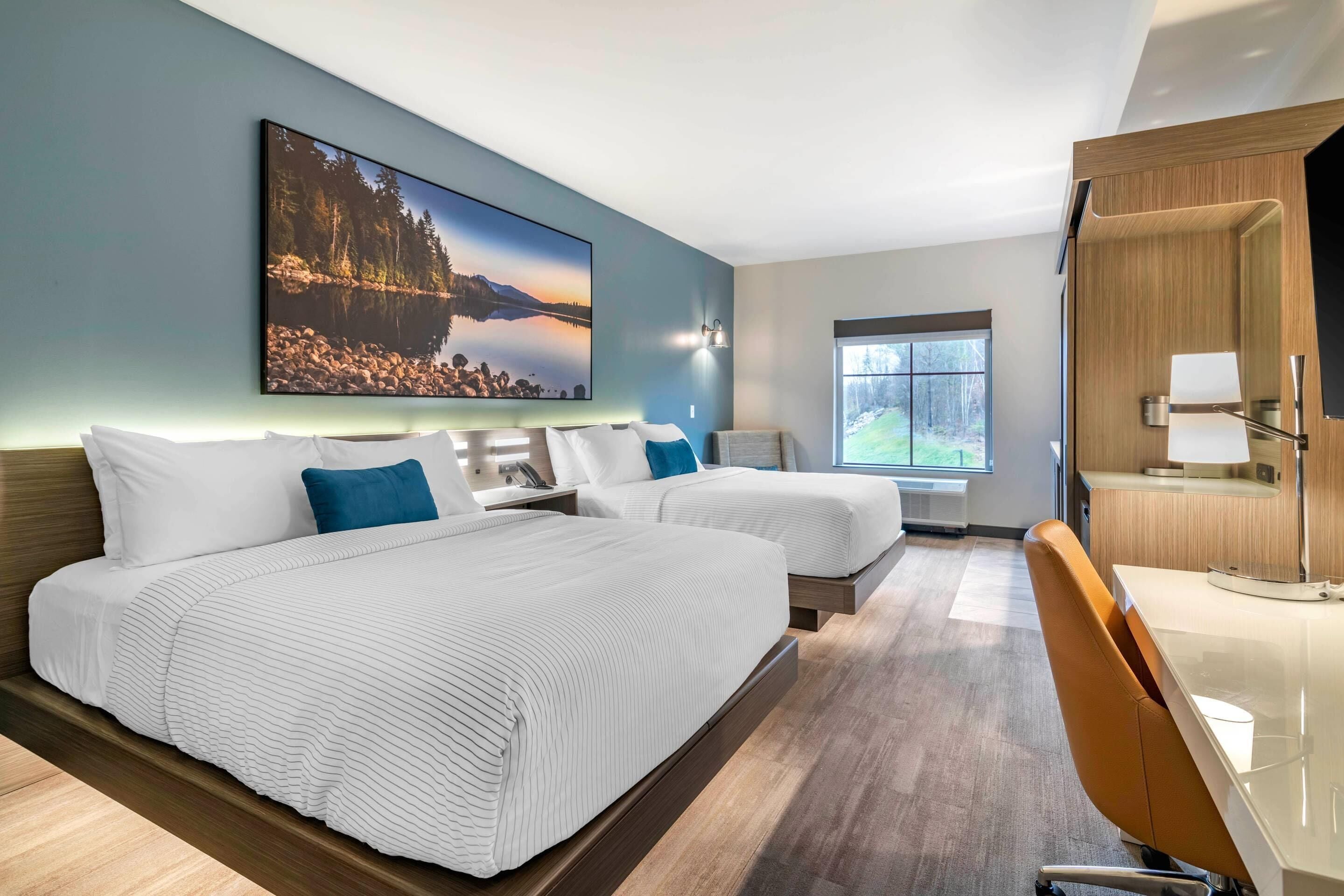 Quarto de hotel com duas camas, uma escrivaninha, uma cadeira, uma janela grande e um quadro na parede.