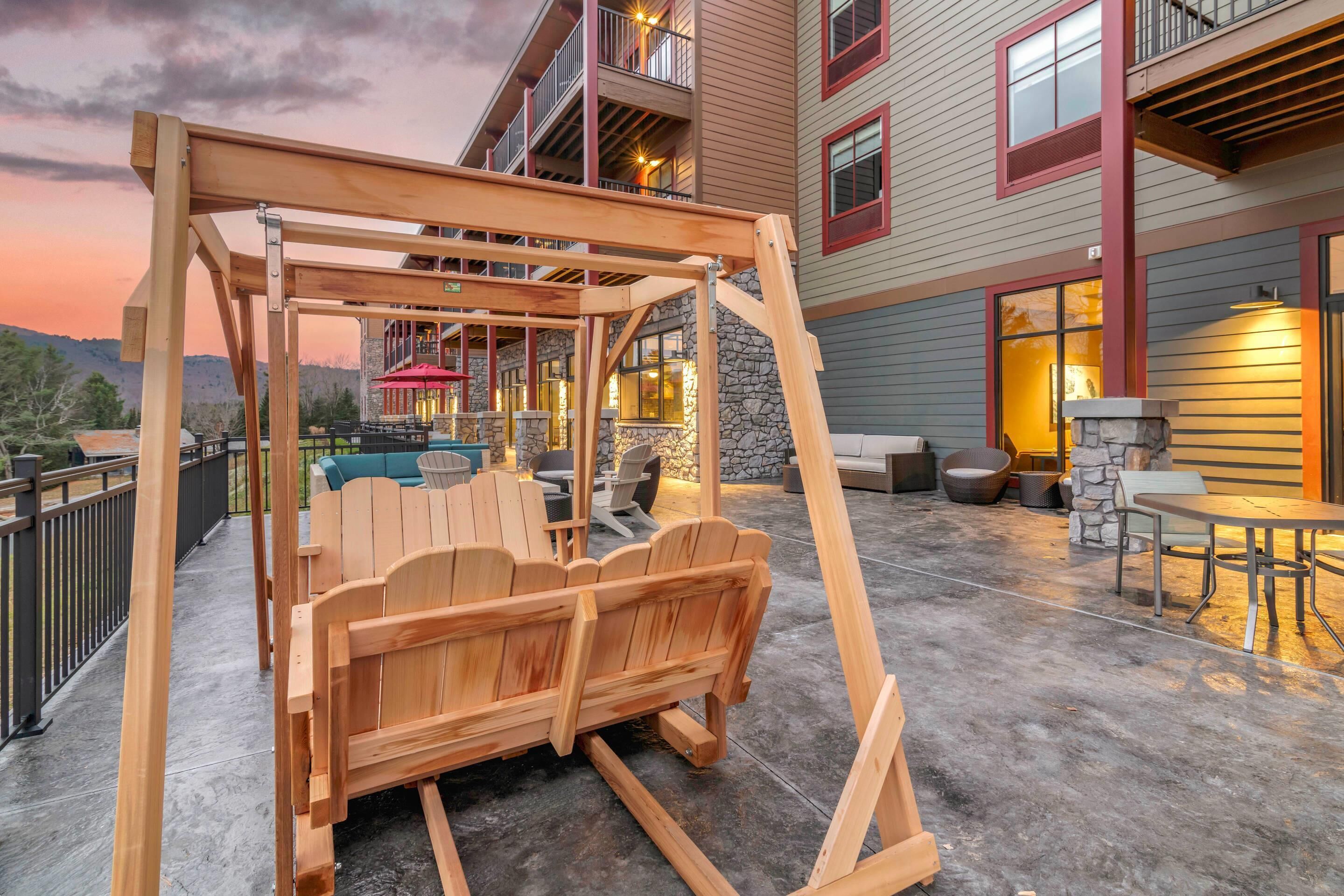 A wooden swing set and seating area on a concrete patio with a view of a building with balconies and windows.