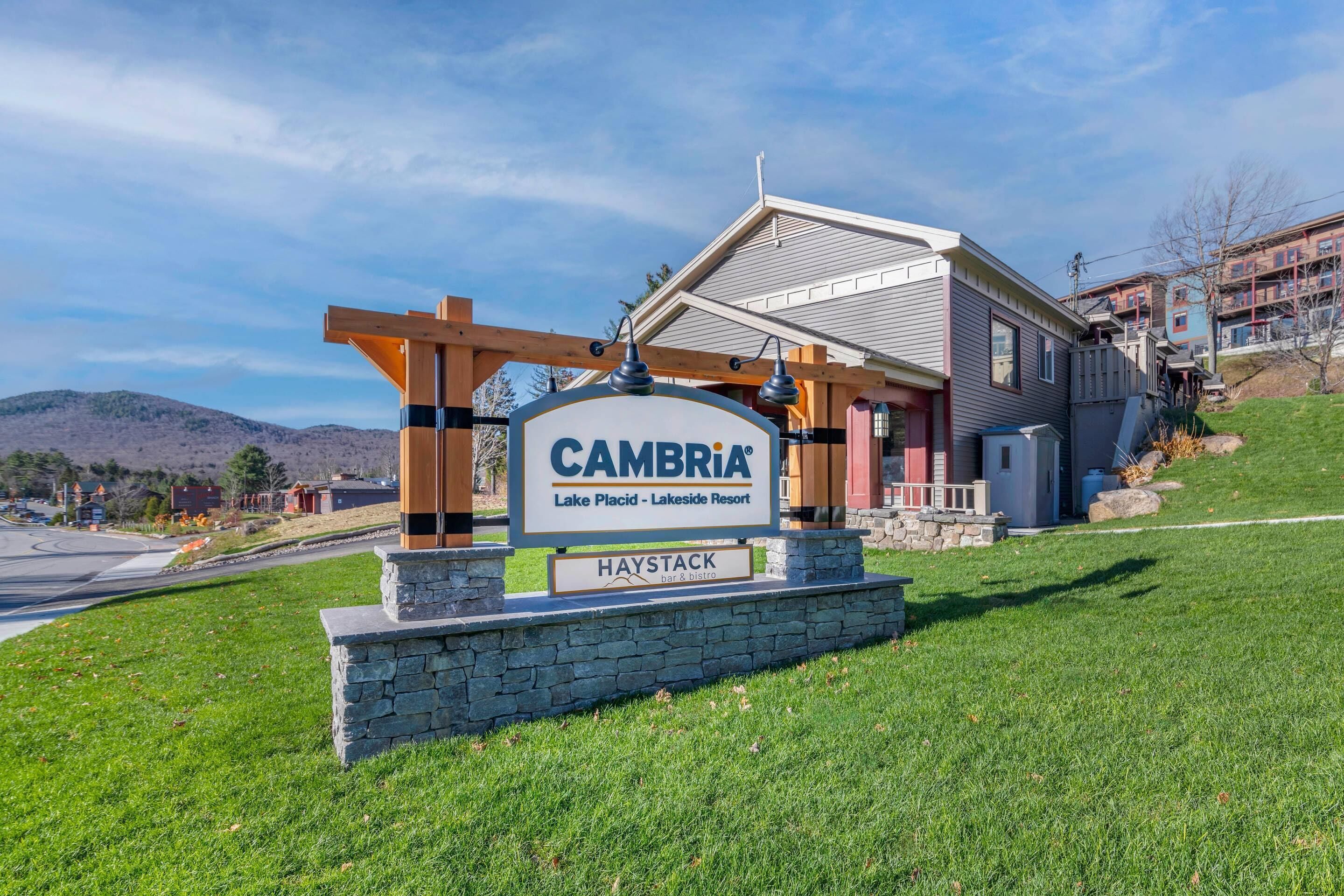 A sign for CAMBRIA Lake Placid - Lakeside Resort with a stone base and a wooden pergola structure.