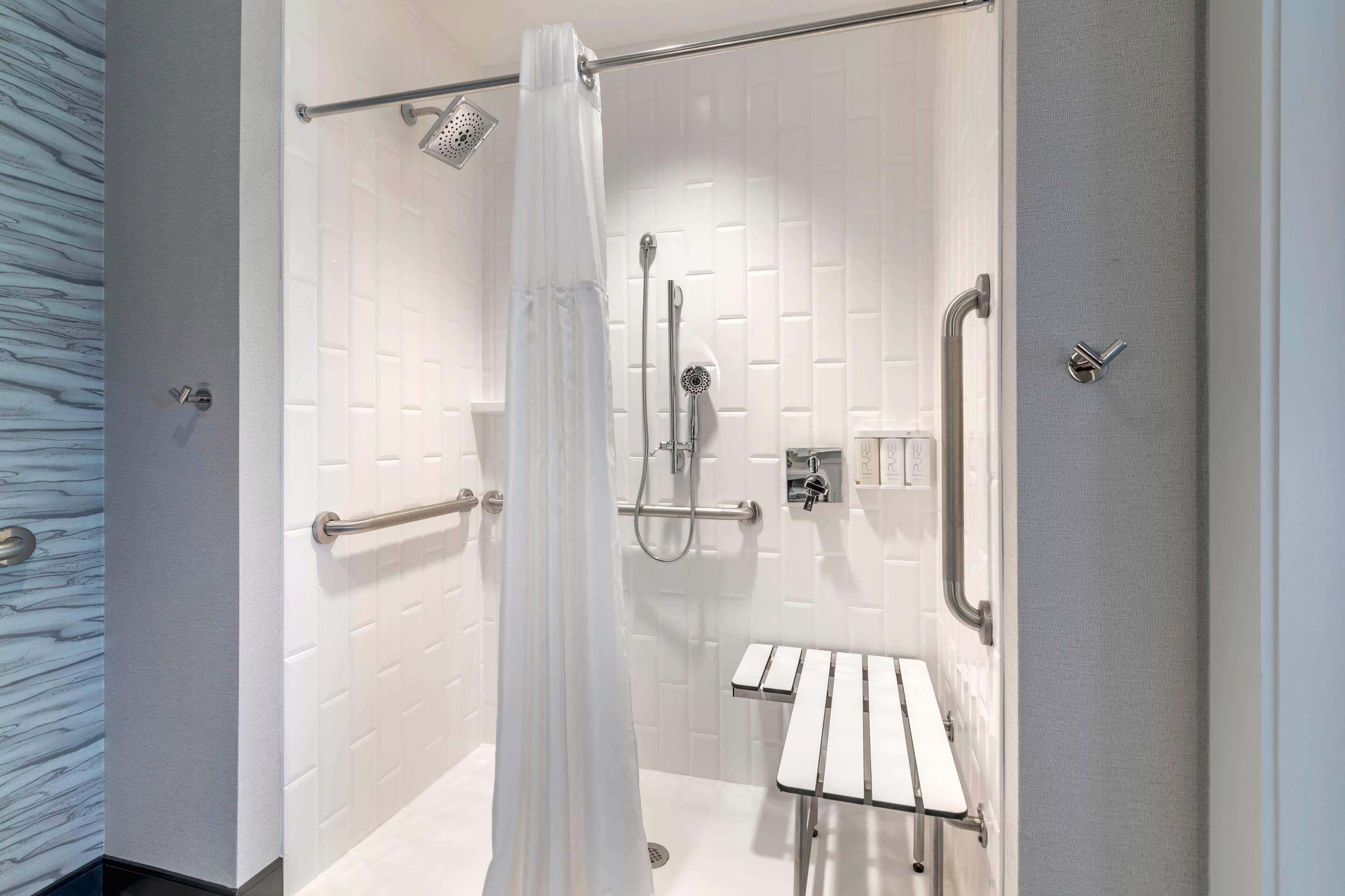 A modern shower area with a white tiled wall, a glass door, a showerhead, a handheld shower, a grab bar, and a small bench.
