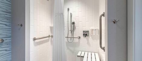 A modern shower area with a white tiled wall, a glass door, a showerhead, a handheld shower, a grab bar, and a small bench.