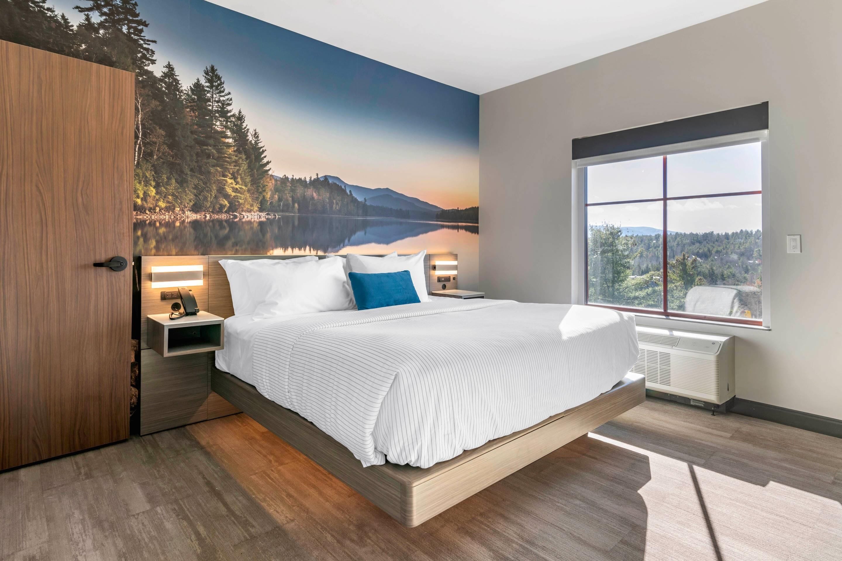 A modern bedroom with a large bed, a nightstand, a wall-mounted lamp, and a window with a view of a lake and mountains.