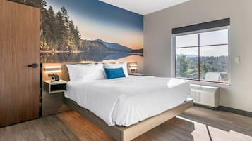 A modern bedroom with a large bed, a nightstand, a wall-mounted lamp, and a window with a view of a lake and mountains.