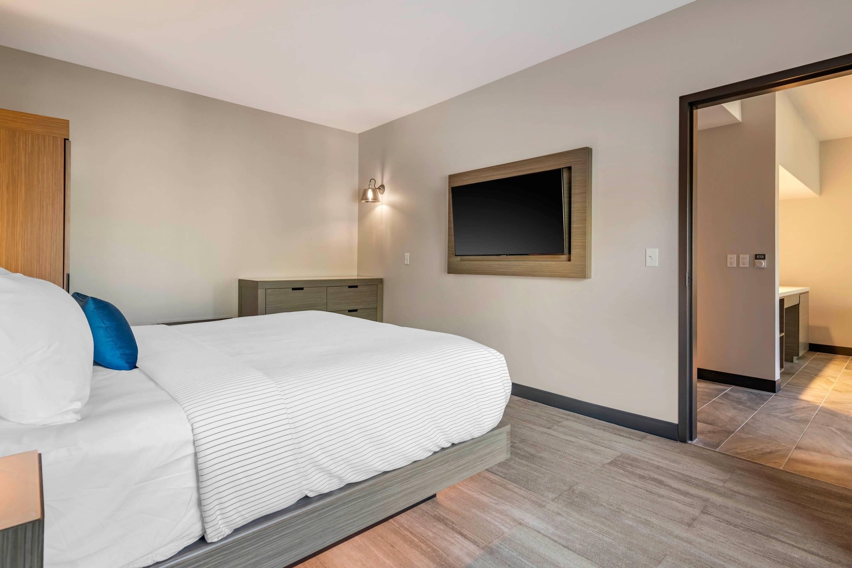 Une chambre d’hôtel moderne équipée d’un lit, d’une table de chevet, d’un téléviseur fixé au mur et d’une armoire en bois.