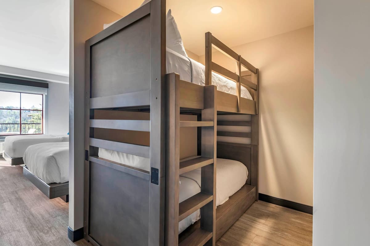 A modern hotel room with a bunk bed, a separate bed, and a large window offering a city view.