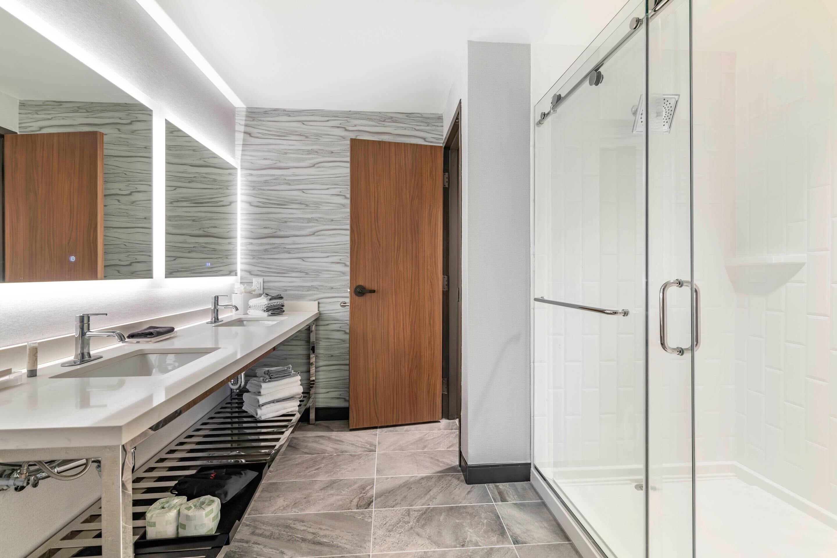 A modern bathroom with a double sink vanity, a glass shower enclosure, and a marble-patterned wall.