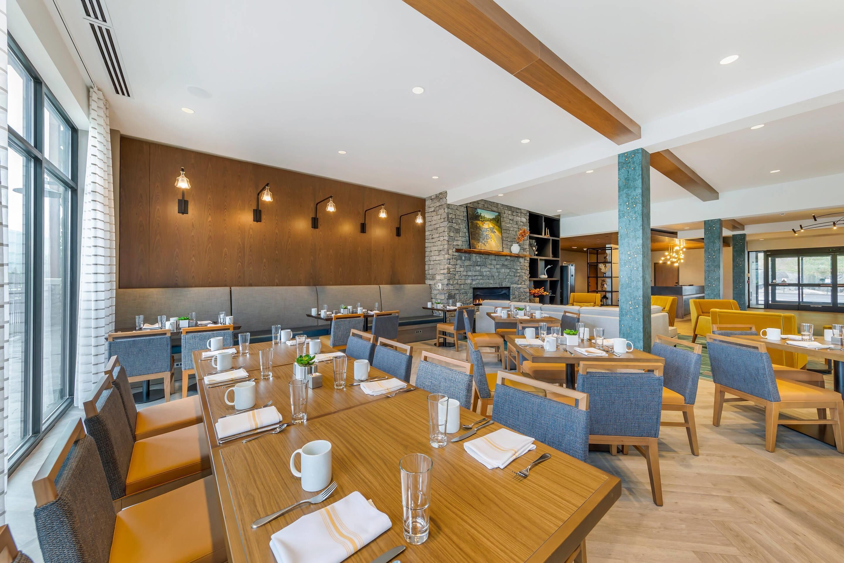 Un restaurant moderne avec des tables et des chaises en bois, une cheminée en pierre et une grande fenêtre donnant sur la ville.