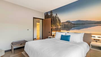 Una habitación de hotel moderna con una cama grande, mesitas de noche, un pequeño rincón de descanso y un mural con vistas a un lago y un bosque.