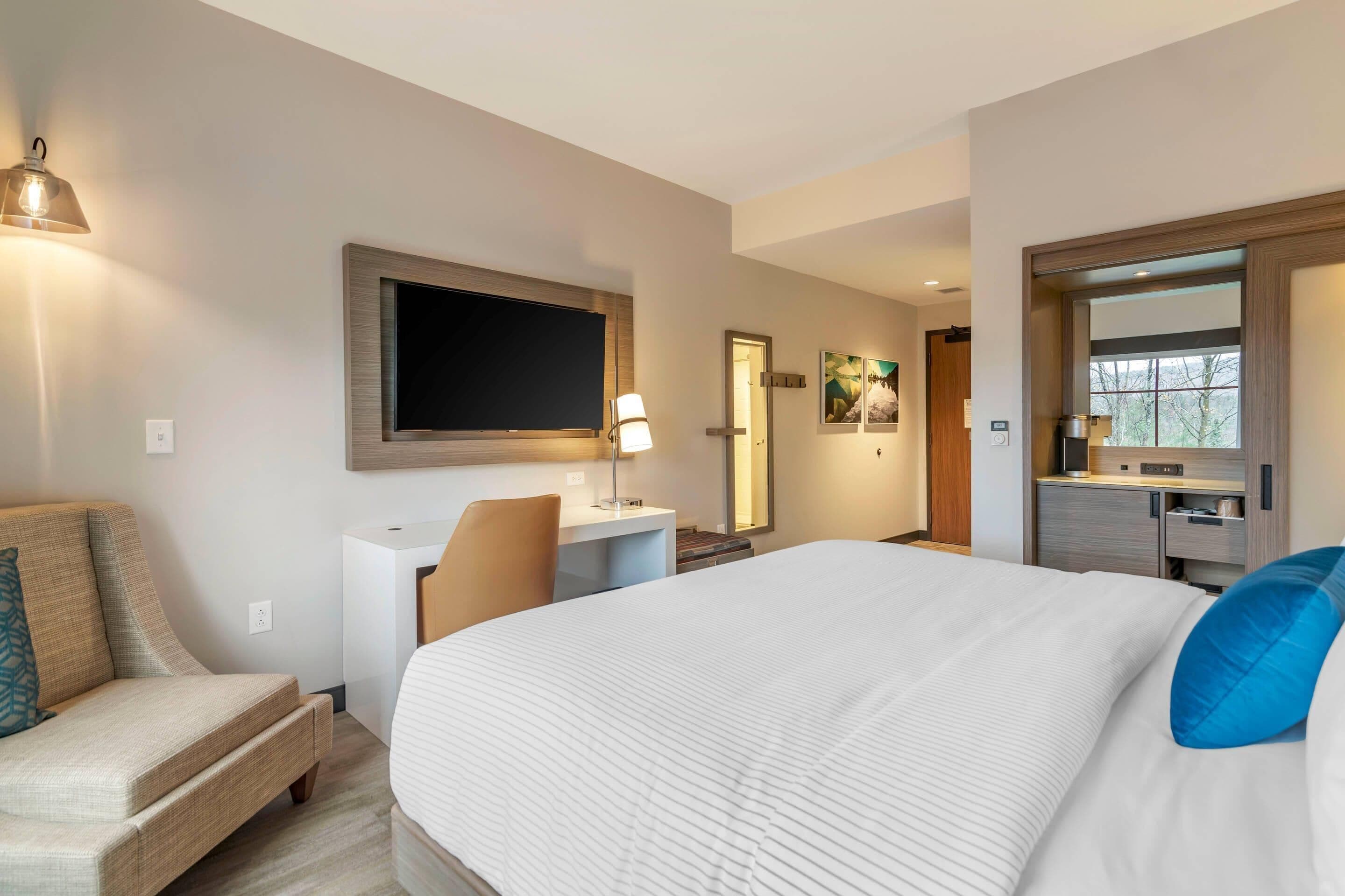 A hotel room with a large bed, a desk, a chair, a TV, and a bathroom visible through an open door.