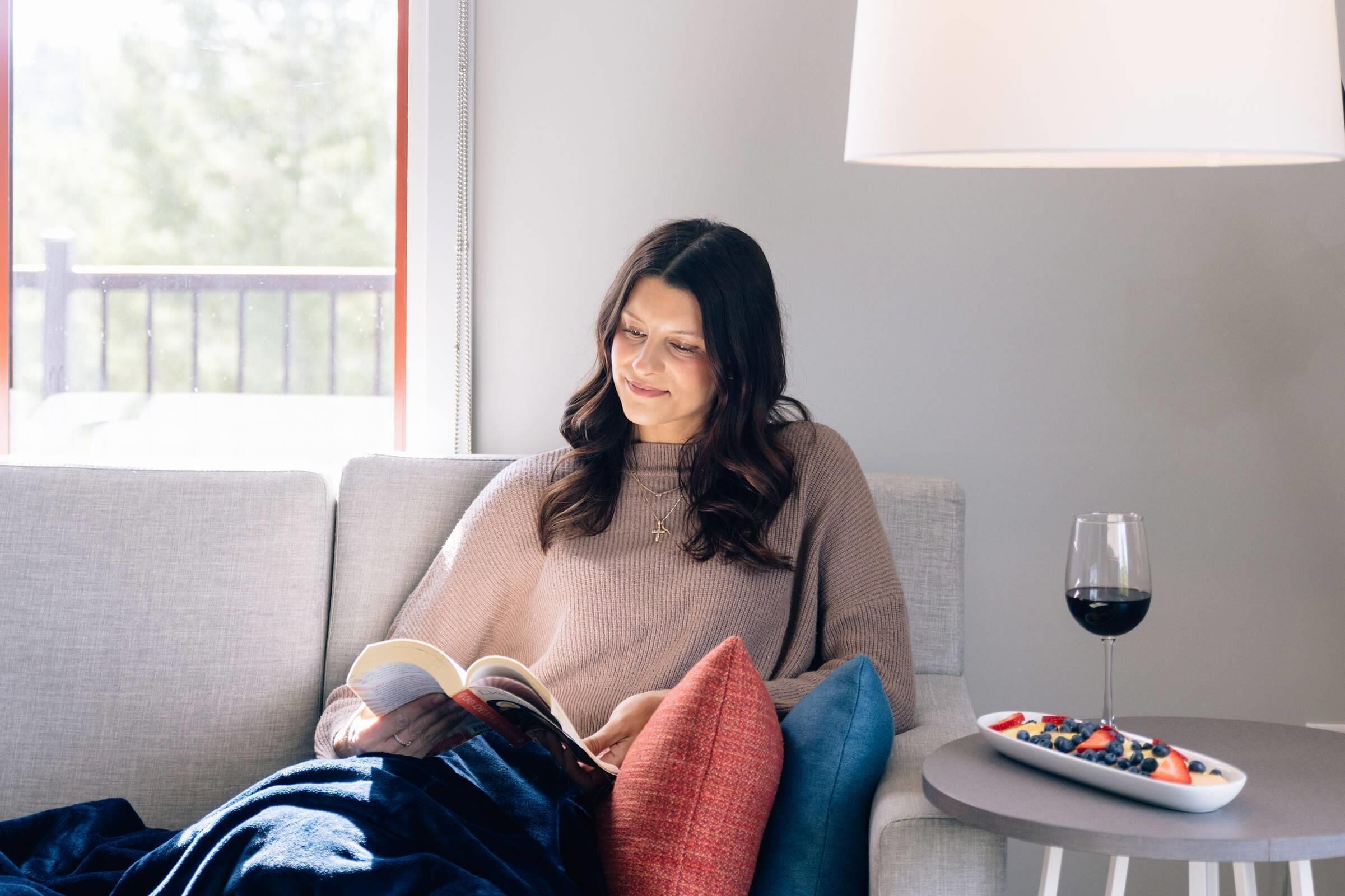 A person sitting on a sofa with a book, a glass of red wine, and a plate of fruit on a small table beside them.
