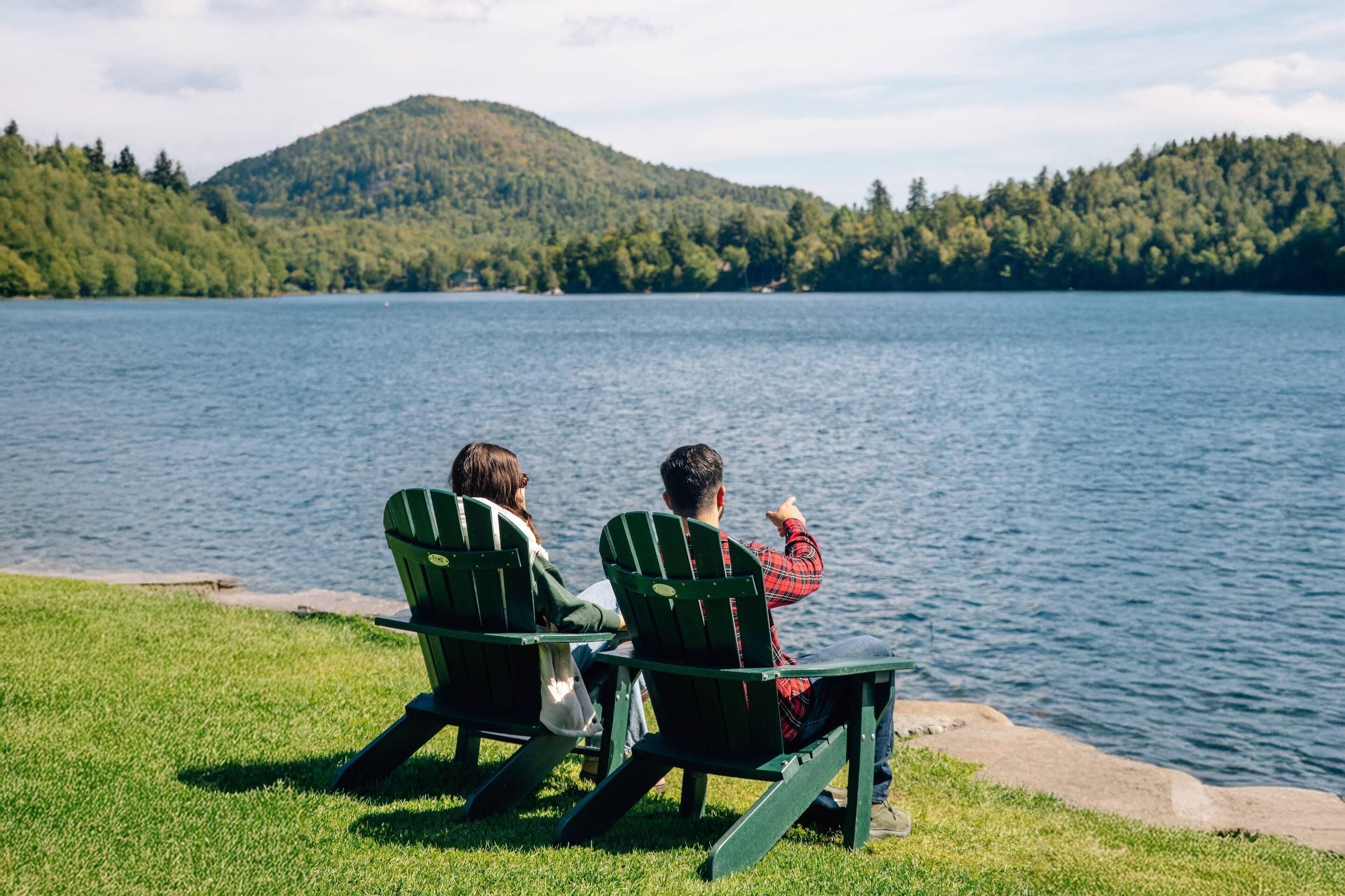 Dos personas sentadas en sillas Adirondack mirando un lago con una colina boscosa al fondo.