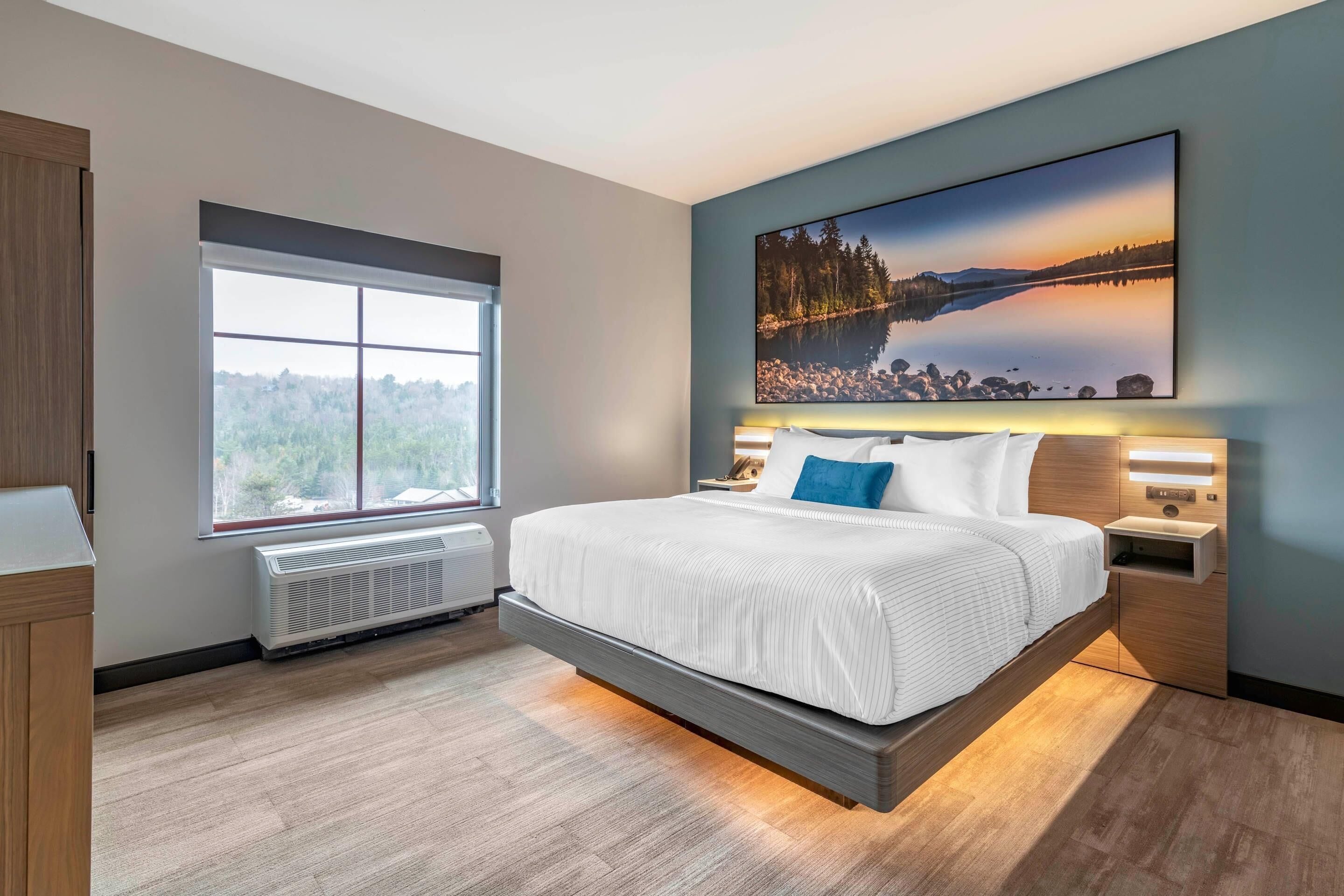 Un dormitorio moderno con una cama grande, un ventanal con vistas a árboles, un cuadro de un lago y un calefactor empotrado.