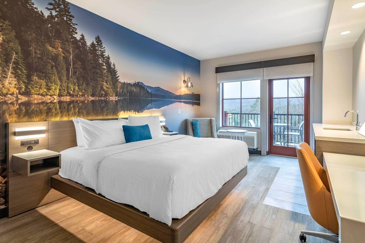A modern hotel room with a large bed, a desk, and a view of a lake through the window.