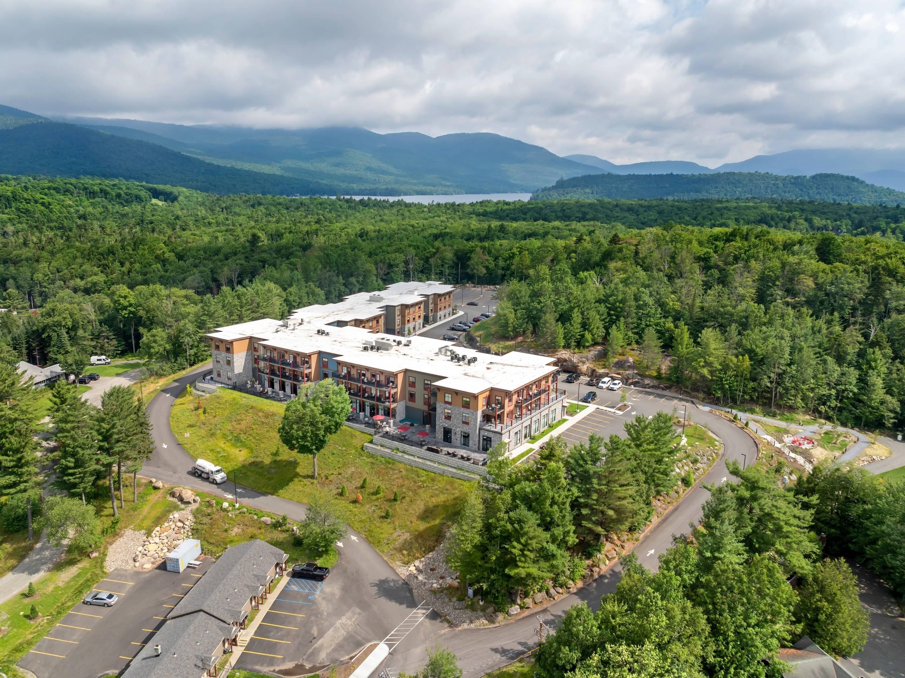 A multi-story building with a parking lot and a road curving around it, surrounded by dense forest and mountains in the background.